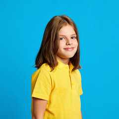 Side view portrait of beautiful teen girl wearing yellow polo shirt, posing with little tender smile on blue studio background. Happy confident child. Concept of childhood, kids emotions, lifestyle