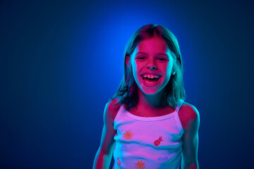 Portrait of young girl emotionally posing, cheerfully laughing and looking at camera against blue studio background in neon light. Happiness. Concept of childhood, kids emotions, lifestyle