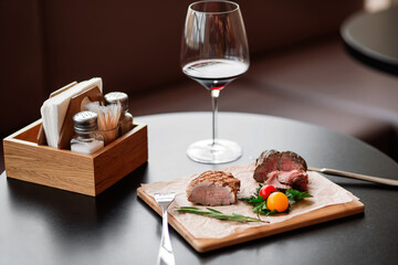 Slices of cooked meat on a wooden cutting board with garnishes of green herbs and cherry tomatoes. A wooden tray with salt, pepper shakers, and a napkin holder is nearby. natural light. 