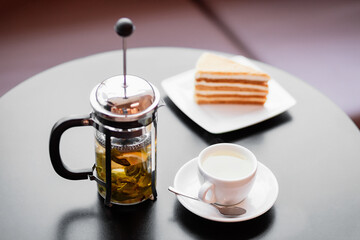 A French press with tea, a white cup partially filled with tea, and a plate with a slice of layered cake on a table. The setting is indoors with natural light. Close-up shot.