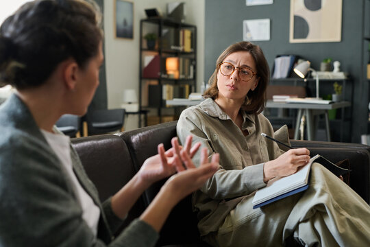 Two women sitting on couch discussing treatment plan during counseling session in cozy office environment with papers and pen in hand