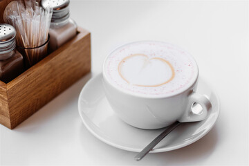White cup of coffee with heart-shaped foam design on a saucer with a spoon. Background has a wooden holder with sugar packets and condiments. Close-up shot on a white surface.
