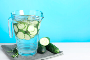 Refreshing cucumber water with rosemary in jug and vegetables on white table against light blue background. Space for text