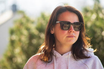 A YOUNG beautiful dark-haired girl in a hoodie stands seriously in sunglasses in the backyard of her house. Difficult life concept