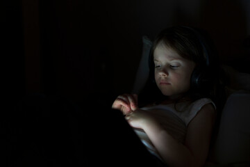 A small beautiful girl wearing headphones lies with a tablet in a dark room. The concept of modern digital life and childhood