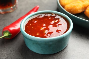 Hot chili sauce in bowl and pepper on grey textured table, closeup