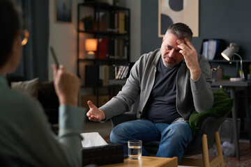 Hispanic man sitting in office talking to professional therapist while holding head in hand,...