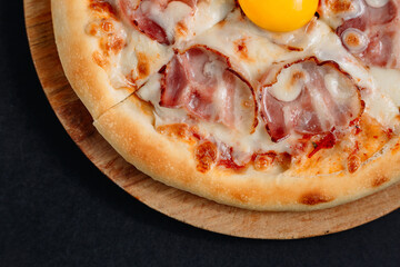 Pizza Carbonara with various meats, melted cheese, and a single egg in the center on a wooden board on a dark surface top view close-up.
