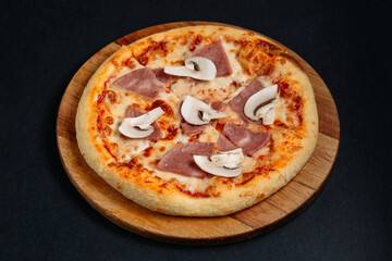 Freshly baked pizza with ham and mushrooms on wooden board on black background