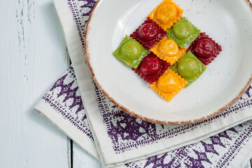  A plate of colorful stuffed pasta green, yellow, red on a patterned napkin. The dish is likely Italian ravioli. The close-up shot emphasizes the intricate details of the pasta.