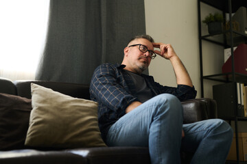 Man relaxing on couch in living room, resting head on hand while considering thoughts with serious...