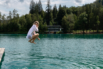 a girl jumps from a wooden pier into the turquoise water of Lake Bled. The child is wearing a white shirt. summer vacation concept, relaxation by the water. Children having fun