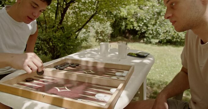Two friends enjoy playing backgammon outdoors on a sunny day. They are seated at a table with coffee cups.