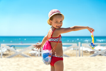 Little girl in swimsuit and hat with bag with sand toys and swimming goggles looks at camera and laughs on sandy beach with blue sea. Vacation summer travel holiday