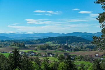 Fototapeta premium Alpine landscape with church in small village Stein im Jauntal nestled amidst rolling hills and lush greenery. Majestic mountain range with snow-capped peaks in Carinthia. Breathtaking backdrop