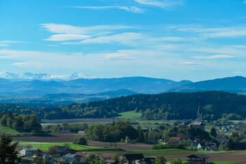 Fototapeta premium Alpine landscape with church in small village Stein im Jauntal nestled amidst rolling hills and lush greenery. Majestic mountain range with snow-capped peaks in Carinthia. Breathtaking backdrop