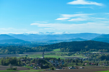 Fototapeta premium Alpine landscape with church in small village Stein im Jauntal nestled amidst rolling hills and lush greenery. Majestic mountain range with snow-capped peaks in Carinthia. Breathtaking backdrop