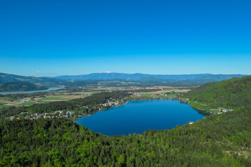 Breathtaking aerial view of a tranquil lake Klopein nestled amidst lush green forest in Carinthia, Austria. Crystal-clear water reflects vibrant blue sky. Rolling hills and mountains in Austrian Alps
