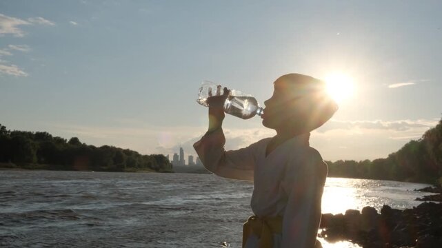 A boy drinks water from a bottle. Silhouette against the background of the river and the city.