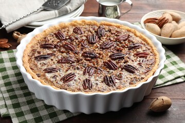 Delicious pecan pie in baking dish and fresh nuts on wooden table, closeup