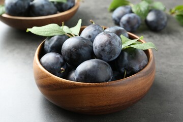 Tasty ripe plums and leaves in bowl on grey table, closeup