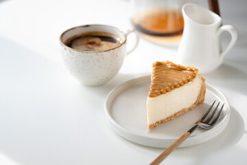 Slice of cheesecake with caramel sauce topping and cup of black american coffee. White table background.