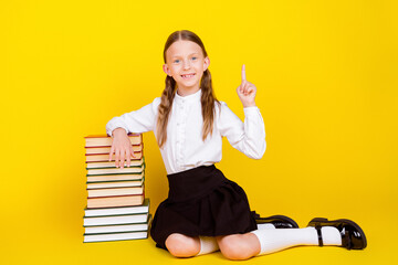 Full length photo of lovely small schoolgirl sit floor book pile point up empty space dressed uniform isolated on yellow color background