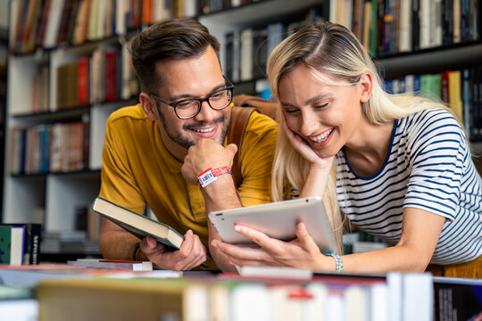 A cheerful Caucasian male and female student discuss academic content using a digital tablet and book in a library setting, donning casual attire.
