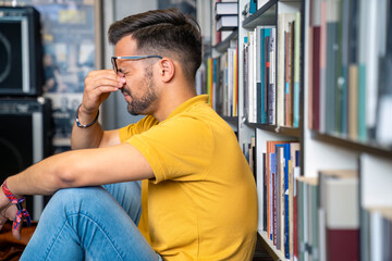 A young Caucasian man in yellow shirt and glasses exhibits stress, sitting on the floor amidst library aisles, with books as his backdrop.