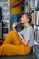 A happy Caucasian female student in casual attire sits on the floor of a library, engrossed in a...
