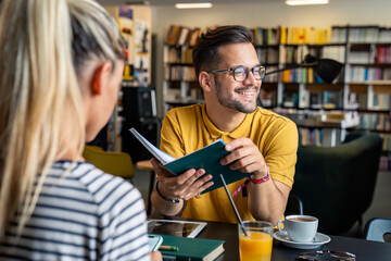 A joyful Caucasian male student holding a book converses with a female peer over refreshments,...