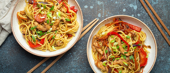 Two bowls with Chow Mein or Lo Mein, traditional Chinese stir fry noodles with meat and vegetables, served with chopsticks top view on rustic blue concrete background.