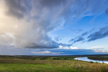Clouds gather above a glistening river, as the sun sets over lush green fields, creating a serene and picturesque twilight atmosphere.