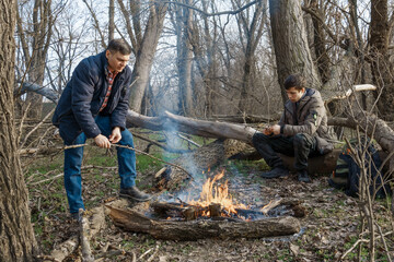 Two men make a campfire for cooking in the forest, father and son sitting on a log while hiking and outdoor activities, early spring landscape