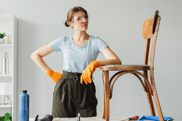Confident carpenter woman wearing protective glasses and gloves posing with old wooden chair in workshop