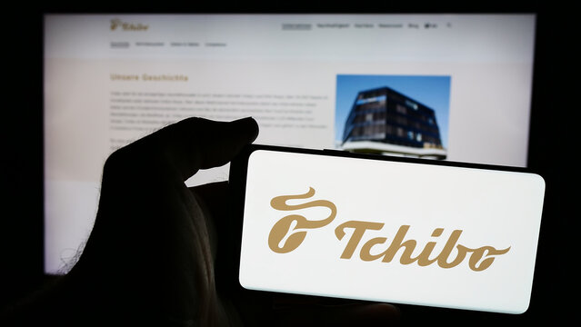 Stuttgart, Germany - 08-07-2024: Person holding mobile phone with logo of German coffee retail company Tchibo GmbH in front of business web page. Focus on phone display.
