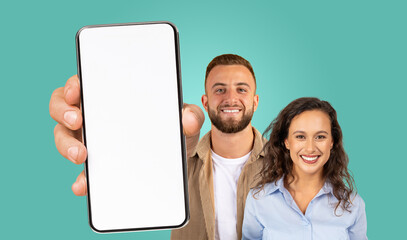 A man and a woman are standing together against a teal background. The man is holding a smartphone with a blank screen towards the camera. They are both smiling and looking directly at the camera.
