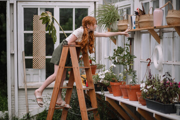 Cute red-haired teenage girl takes care of and plants plants in a greenhouse. On the stairs, she pulls out a book on gardening in the spring for Earth Day.