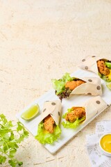 Mini tortilla wraps filled with fried chicken, cheese, salad and mustard sauce on a ceramic board on a light concrete background.