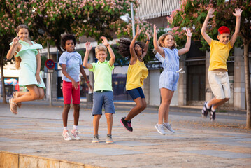 Obraz premium Multiracial group of cheerful emotional preteen friends gaily spending time on summer day, jumping together on city street..