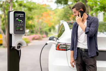 Man talking on smartphone while recharging electric car battery charging from EV charging station...