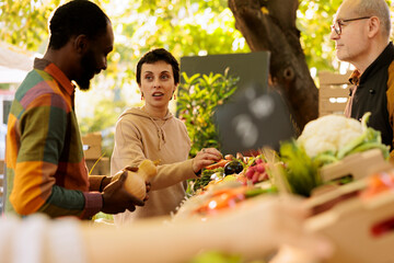 Elderly small farm business owner helping diverse customers with purchasing locally grown fruits and vegetables. Multiracial couple buying fresh organic bio food products from senior vendor.
