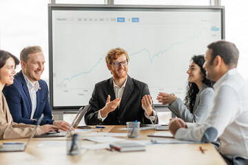 A group of diverse business professionals are gathered around a conference table in a modern office setting. They are looking at large screen displaying a growth chart, engaging in lively discussion