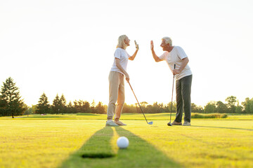 An elderly couple in sportswear exchange greetings on a golf course after a successful shot. Man...