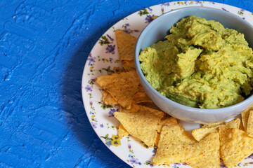 Guacamole in a blue bowl surrounded by nachos on a plate decorated with flowers on a blue background