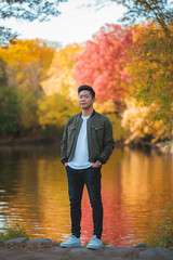 Fototapeta premium A young man standing by a lake surrounded by vibrant autumn foliage. He looks serene, blending in with the natural beauty around him, representing peacefulness and a deep connection with nature.