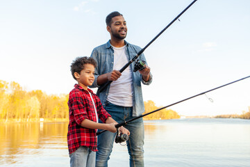 An African American father and son stand on a wooden pier near a lake holding fishing rods. An...