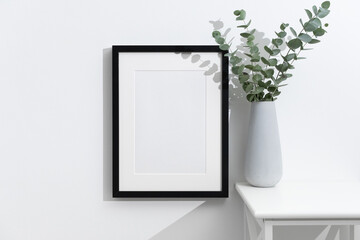 Vertical black artwork frame mockup on white wall with fresh eucalyptus bouquet in big grey vase, blank mock up with copy space