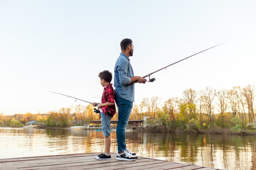 An African American father and his son stand back to back on a wooden pier by a lake, holding fishing rods. The father is wearing a denim jacket and jeans, the son is wearing a red plaid shirt and