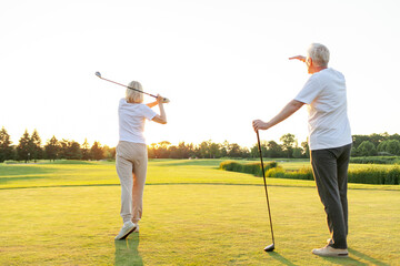 An elderly couple in sports clothes playing golf on a field. The woman stands in the distance swinging a club, the man behind her back watching the shot The bright sun complements the atmosphere. 